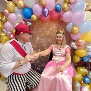 Kobieta w różowej sukni księżniczki i mężczyzna w kostiumie pirata siedzą w otoczeniu kolorowych balonów, pozując na tle błyszczącej dekoracji.