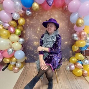 Kobieta w eleganckiej sukience w fioletowych odcieniach z szaro-białym szalem, siedząca na krześle przed tłem ozdobionym kolorowymi balonami i błyszczącymi akcentami, idealna na przyjęcie lub imprezę tematyczną.