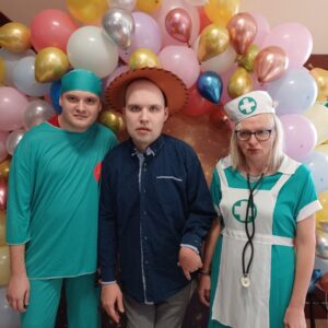Grupa trzech osób w kostiumach medycznych pozująca przed balonową dekoracją. Dwie osoby w strojach lekarzy: jedna w zielonym uniformie, a druga w białej sukience pielęgniarki z stetoskopem. Trzecia osoba w niebieskiej koszuli z brązowym kapeluszem.