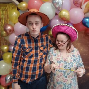 Młoda para pozująca na imprezie w kolorowych kapeluszach przed tłem z balonami. Mężczyzna w koszuli w kratę i kobieta w sukience w liście trzymają żółtą wstążkę.