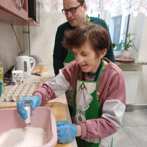 Osoba używająca soli do przygotowania ciasta w kuchni, z mężczyzną w tle. Obie osoby noszą fartuchy i rękawice, a na blacie widoczne są składniki do pieczenia.