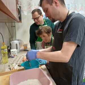 Osoby przygotowujące składniki do pieczenia w kuchni, z widoczną miską mąki i jajkiem. W tle różne przybory kuchenne, a uczestnicy noszą fartuchy i rękawice.