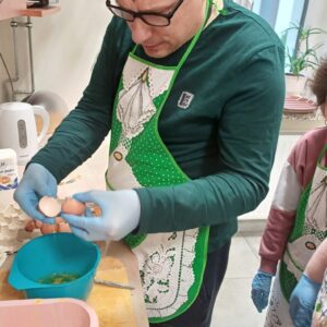 Mężczyzna w zielonym fartuchu rozbija jajka do miski podczas gotowania w kuchni. Obok niego stoi dziecko w różowym swetrze, obydwoje mają założone rękawice ochronne. W tle widoczne akcesoria kuchenne oraz składniki do pieczenia.