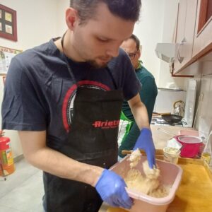 Mężczyzna przygotowujący ciasto w kuchni, noszący czarny fartuch i niebieskie rękawice, mieszający składniki w różowym pojemniku. W tle widać drugą osobę i przybory kuchenne.