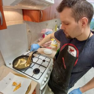 Mężczyzna gotuje w kuchni, używając szczypiec do wyciągania potrawy z patelni. W tle widoczna jest kuchenka gazowa oraz blaty kuchenne z różnymi akcesoriami. Mężczyzna ma na sobie czarny fartuch i rękawice, a na stole leżą usmażone potrawy.