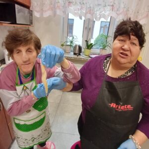 Dwie kobiety w kuchni trzymają cienki kawałek ciasta. Obie noszą rękawice ochronne. W tle widoczne są rośliny doniczkowe na parapecie oraz sprzęt kuchenny.