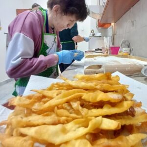 Kobieta w fartuchu przygotowuje potrawy w kuchni, koncentrując się na nawijaniu ciasta, podczas gdy w tle widoczne są złociste chrupiące smakołyki na papierze kuchennym.