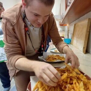Mężczyzna w jasnym ubraniu układa chrupiące frytki na talerzu w kuchni. W tle widać inne osoby oraz akcesoria kuchenne. Frytki są złociste i apetyczne, co sugeruje świeże przygotowanie.