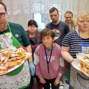 Grupa osób w kuchni, trzymająca talerze z domowymi faworkami, posypanymi cukrem pudrem. W scence widoczna radosna atmosfera w trakcie wspólnego gotowania.