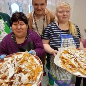 Osoby trzymające talerze z pysznymi, posypanymi cukrem pudrem faworkami, w przyjaznej atmosferze kuchennej.