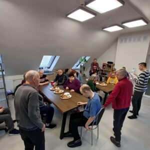 Grupa osób starszych zasiadająca przy stole w jasnym pomieszczeniu, gdzie zjedzą posiłek. Na stole talerze z jedzeniem, a w tle widać rośliny oraz inni uczestnicy spotkania.
