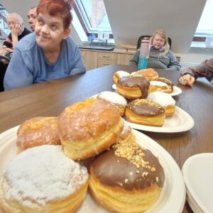 Stół w jadalni z talerzami pełnymi pączków, w tym pączki w czekoladzie i posypce orzechowej, podczas spotkania towarzyskiego. W tle widać kilka osób, w tym dwie osoby siedzące i rozmawiające.