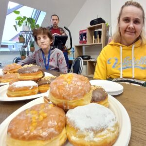 W tle dwóch osób siedzących przy stole, na pierwszym planie znajduje się talerz z różnorodnymi pączkami, w tym pączki z lukrem, posypką orzechową i owocami. Scena odbywa się w jasnym pomieszczeniu z roślinami doniczkowymi i sprzętem do ćwiczeń.