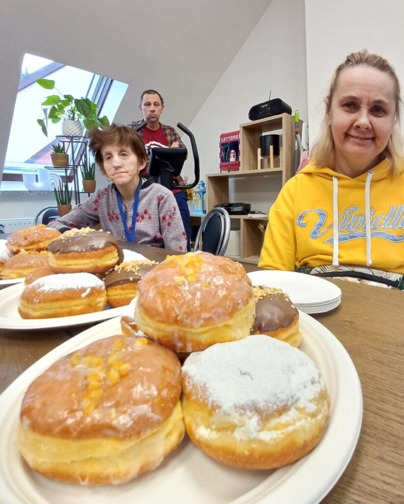 W tle dwóch osób siedzących przy stole, na pierwszym planie znajduje się talerz z różnorodnymi pączkami, w tym pączki z lukrem, posypką orzechową i owocami. Scena odbywa się w jasnym pomieszczeniu z roślinami doniczkowymi i sprzętem do ćwiczeń.