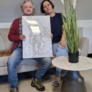 Para siedząca na kanapie, trzymająca rysunek przedstawiający postać w górach. W tle widać dekoracyjną roślinę w doniczce oraz nowoczesny stół.