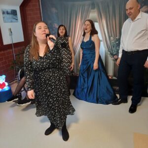 Kobieta w czarnej sukience z białymi wzorami śpiewa do mikrofonu, otoczona przez trzy osoby. Dwie z nich są w eleganckich strojach, jedna w sukience wieczorowej, a druga w sukience w paski. Tło przedstawia zasłony i kwiaty, co nadaje przyjemny klimat. Scena uchwycona w trakcie imprezy lub występu.