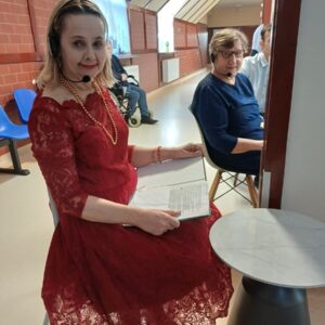 Kobieta w czerwonej, koronkowej sukience siedzi przy stole, trzymając notatki. Ma na sobie słuchawki i makijaż, uśmiecha się do kamery. W tle widać innych uczestników wydarzenia oraz krzesła. Pomieszczenie ma jasne ściany i dużą ilość naturalnego światła.