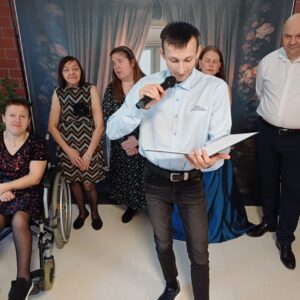 Grupa osób na wydarzeniu społecznym, w tym mężczyzna trzymający mikrofon i czytający, oraz cztery kobiety, z których jedna jest na wózku inwalidzkim. Tło stanowi elegancka zasłona i dekoracyjne elementy.