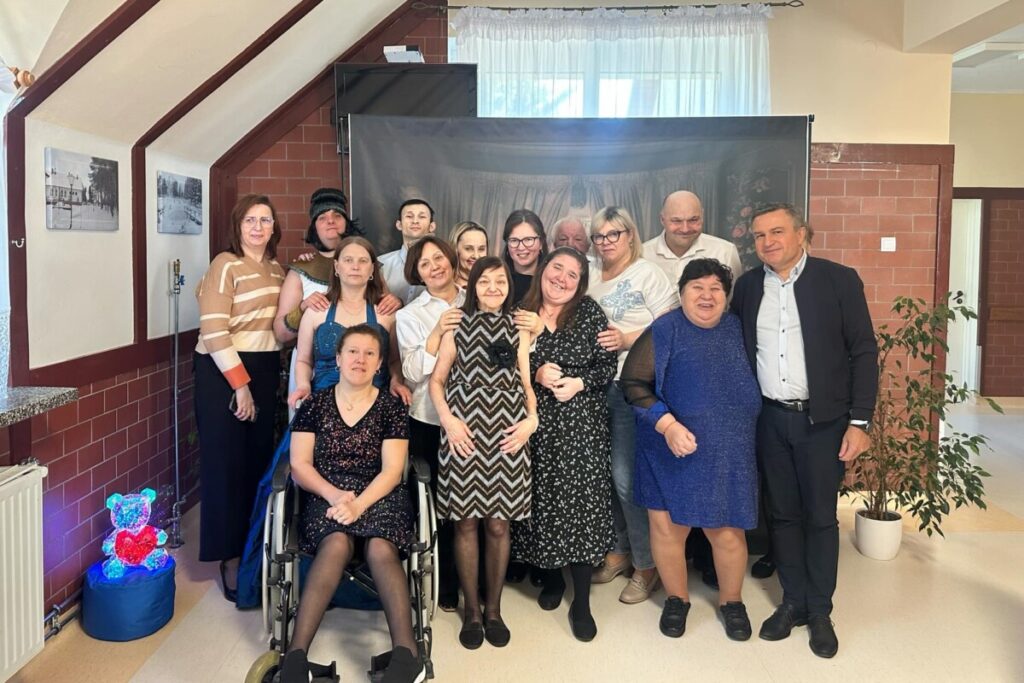 Grupa osób pozująca do zdjęcia w placówce wsparcia społecznego, w tle zdjęcia historyczne, dekoracje w postaci świecącego misia. Wszyscy uśmiechnięci, reprezentujący różne style ubioru.