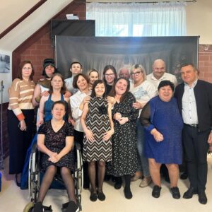 Grupa osób pozująca do zdjęcia w placówce wsparcia społecznego, w tle zdjęcia historyczne, dekoracje w postaci świecącego misia. Wszyscy uśmiechnięci, reprezentujący różne style ubioru.