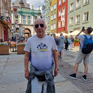 Starszy mężczyzna w okularach przeciwsłonecznych, ubrany w t-shirt z napisem "California", stoi na malowniczej ulicy pełnej kolorowych kamienic. W tle widać tłum ludzi oraz zabytkowy budynek. Słoneczny dzień sprzyja spacerom i działalności turystycznej.