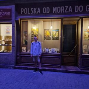 Młody mężczyzna stoi przed sklepem z latarniami, w tle wystawa z plakatami przedstawiającymi Polskę. Na witrynie widoczne są książki i foldery o tematyce turystycznej oraz historycznej, a atmosfera jest wieczorna, z ciepłym oświetleniem.