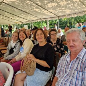 Grupa osób siedzących pod namiotem na wydarzeniu plenerowym, uśmiechnięte twarze kobiet i mężczyzn, w tle widoczne krzesła i uczestnicy wydarzenia.