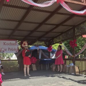 Kobiety w czerwonych sukienkach występują na scenie w dekorowanej altanie, podczas wydarzenia z hasłem "Kocham Cię Polsko". W tle widoczne są kwiaty i prezenty, co tworzy radosną atmosferę.