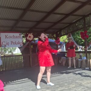 Kobieta w czerwonej sukience występuje na scenie podczas wydarzenia kulturalnego, trzyma mikrofon i śpiewa. W tle widoczna jest dekoracja z napisem "Kocham Cię Polsko" oraz inne osoby w strojach festiwalowych.