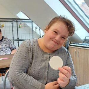 Dziewczyna uśmiechająca się, trzymająca okrągły biały papier w dłoni, siedzi przy stole w jasnym pomieszczeniu. W tle widać inne osoby oraz materiały do rysowania, co sugeruje aktywność artystyczną lub kreatywną.