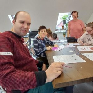 Grupa osób dorosłych uczestniczy w zajęciach plastycznych w jasnym pomieszczeniu. W centrum uwagi znajduje się mężczyzna z krótkimi włosami, który uśmiecha się do kamery, oraz kobieta w paski, pracująca nad rysunkiem. Na stole leżą kolorowe kartki i przybory do rysowania. W tle widać inne osoby skupione na twórczości artystycz
