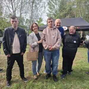 Grupa pięciu osób pozujących na tle zielonego terenu, w otoczeniu drzew. Na zdjęciu widoczni mężczyźni i kobieta, ubrani w casualowe stroje, z uśmiechami na twarzach. W tle czarny namiot, sugerujący wydarzenie plenerowe.