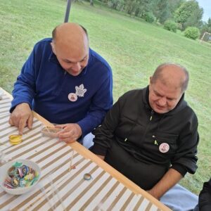 Dwóch mężczyzn siedzi przy stole na zewnątrz, zajętych kreatywnym projektem. Na stole znajdują się różne materiały rzemieślnicze, a w tle widać zieleń otaczającego terenu. Obaj mężczyźni mają uśmiechy na twarzach, co sugeruje przyjemną atmosferę.