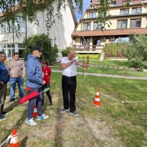 Mężczyzna strzelający z łuku podczas warsztatów łuczniczych, z grupą uczestników w tle, w otoczeniu zieleni i budynków mieszkalnych.
