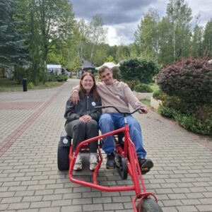 Para młodych ludzi siedząca na czerwonym rowerze trójkołowym na ścieżce w parku, otoczeni zielenią i drzewami. W tle widać chmurzyste niebo.