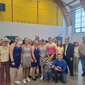 Grupa ludzi w strojach tanecznych, pozująca do zdjęcia w hali sportowej. W tle widać zegar sportowy oraz uczestników wydarzenia. Atmosfera radosna, z różnorodnymi strojami i uśmiechami.