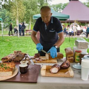 Mężczyzna kroi pieczone mięso na stoisku gastronomicznym na świeżym powietrzu, obok znajdują się różne rodzaje chleba i dodatków. W tle widać gości biorących udział w wydarzeniu na zielonej trawie.