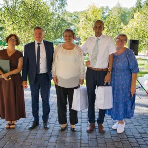 Grupa pięciu osób pozująca do zdjęcia na zewnątrz, w tle zielone drzewa i dekoracje balonowe. Uczestnicy ubrani w eleganckie stroje, trzymają torby. Scena z wydarzenia, prawdopodobnie uroczystości lub nagrody.