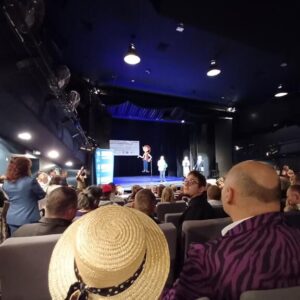 Widok z sali teatralnej podczas wydarzenia, gdzie na scenie występują postacie, a publiczność siedzi w fotelach. Na pierwszym planie widać osobę w kapeluszu słomkowym oraz innych widzów. Scena oświetlona, z tłem w kolorze niebieskim.