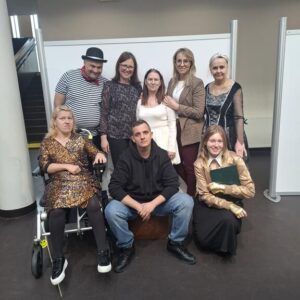 Grupa osób pozująca do zdjęcia w nowoczesnym wnętrzu. Wśród nich znajduje się kobieta na wózku inwalidzkim, a także osoby w różnorodnych stylizacjach, od eleganckich po casualowe. Uśmiechnięte twarze sugerują radosną atmosferę spotkania.