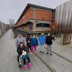 Grupa sześciu osób stoi na chodniku przy starym budynku, obok torów kolejowych. Wszyscy są ubrani w ciepłe kurtki, a jedna osoba siedzi na wózku inwalidzkim. W tle widoczne są szare chmury i nowoczesne elementy architektury.