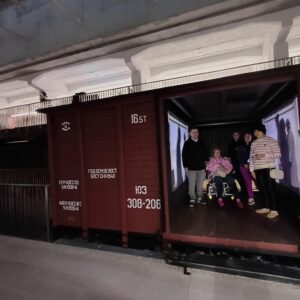 Grupa osób stojąca w wagonie towarowym, z projekcją na ścianach. Wagon ma oznaczenia, w tym numer 165 t. Scena odbywa się w nowoczesnej przestrzeni wystawowej.