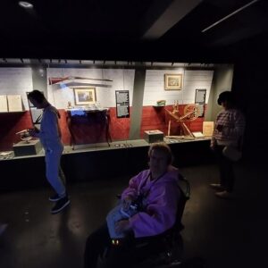 Wystawa muzealna z eksponatami historycznymi, w tym bronią, obrazami i narzędziami rzemieślniczymi. W pierwszym planie osoba na wózku inwalidzkim, w tle zwiedzający podziwiają ekspozycję.