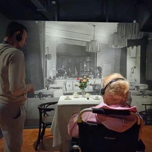 Mężczyzna i starsza kobieta na wózku inwalidzkim w muzeum, stojący przed dużym muralem przedstawiającym historyczną restaurację z kwiatami na stole. Obie postacie słuchają nagrania audio za pomocą słuchawek.