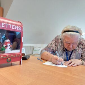 Starsza kobieta pisząca list przy biurku, obok kolorowa skrzynka na listy z motywem świątecznym i bałwankiem.