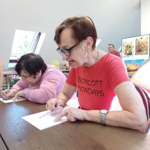 Seniorzy uczestniczą w warsztatach artystycznych, rysując na papierze. Kobieta w czerwonym T-shircie z napisem "BOYCOTT MONDAYS" intensywnie skupia się na swojej pracy, podczas gdy inna uczestniczka, ubrana w różową bluzkę, również rysuje. W tle widać kolorowe prace artystyczne.