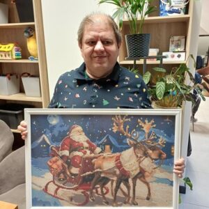 Mężczyzna trzymający obrazek przedstawiający Świętego Mikołaja na saniach ciągniętych przez renifery w zimowej scenerii. W tle widać zaśnieżone domy i gwiazdy na nocnym niebie. Mężczyzna ma na sobie koszulę w świąteczny wzór.