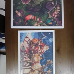 Obrazek przedstawia dwa w ramkach hafty krzyżykowe, umieszczone na drewnianym stole. Górny haft przedstawia kolorowe ptaki w otoczeniu kwiatów, a dolny ukazuje Świętego Mikołaja w saniach ciągniętych przez renifera w zimowej scenerii.