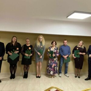 Uczestnicy ceremonii wręczenia nagród, stojący w rzędzie, trzymający zielone dyplomy i róże. W tle jasna ściana, a w sali znajduje się oświetlenie sufitowe.