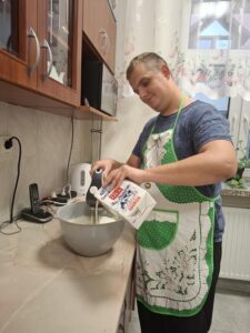 Mężczyzna w fartuchu kuchennym wlewa mleko do miski z ciastem, przygotowując potrawę w kuchni. W tle widoczna jest kuchenne wyposażenie oraz telefon stacjonarny.